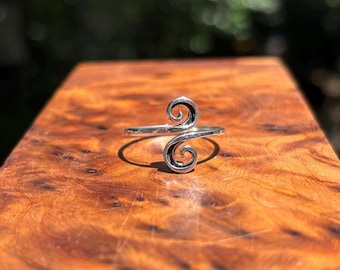 May include: A silver-colored ring with a double swirl design. The ring is set against a wooden surface, with the sunlight reflecting off the polished metal. The ring's design features two opposing spirals.