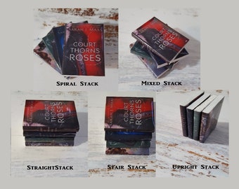 May include: Five different ways to stack a paperback book with the title "A Court of Thorns and Roses" on the cover. The book cover features a red, black, and white design with a woman's face and a rose.