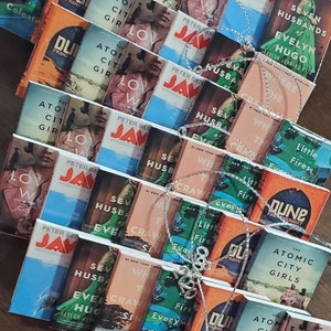 May include: A collection of miniature book ornaments featuring popular novels such as "Dune", "The Atomic City Girls", "Lovely War", "Jaws", "Seven Husbands of Evelyn Hugo", and "Where the Crawdads Sing". The ornaments are silver and feature a variety of colors and designs.