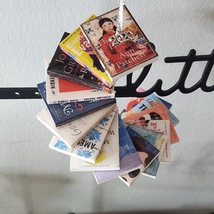 May include: A stack of miniature book ornaments, each with a different book cover design. The top ornament features a book titled "The  House  2020" by Ann Patchett.