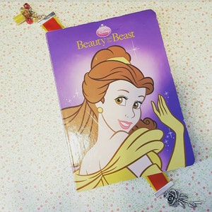 以下が含まれることがあります： 「ディズニー美女と野獣」というタイトルの紫色の本で、黄色のドレスを着たベルのイラストが描かれています。赤い金色のしおりに銀色のチャームが付いています。