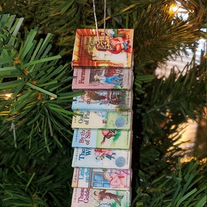 May include: A Christmas ornament featuring a stack of miniature Little House on the Prairie books. The books are arranged vertically and feature colorful illustrations of characters and scenes from the books. The ornament is hung on a silver string.