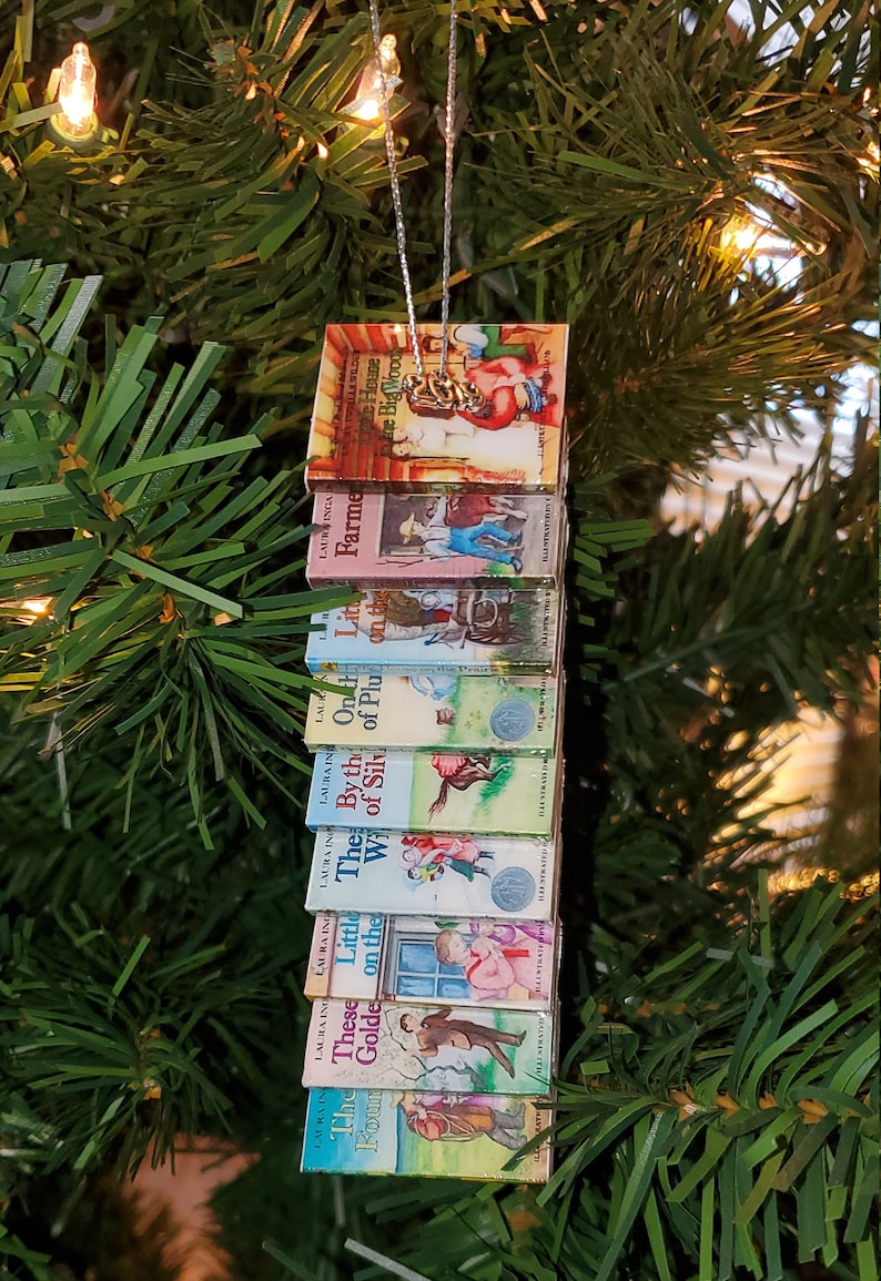 May include: A Christmas ornament featuring a stack of miniature Little House on the Prairie books. The books are arranged vertically and feature colorful illustrations of characters and scenes from the books. The ornament is hung on a silver string.