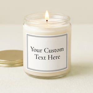 May include: A lit candle in a clear glass jar with a gold-colored metal lid placed to the side. The jar features a white rectangular label with a thin black border and the black printed text Your Custom Text Here centered on the front surface.