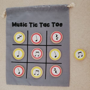 Puede incluir: Una bolsa de tela gris con la inscripci&oacute;n "Music Tic Tac Toe". El juego es un tres en raya con fichas de madera con notas musicales. Una ficha est&aacute; separada del tablero.