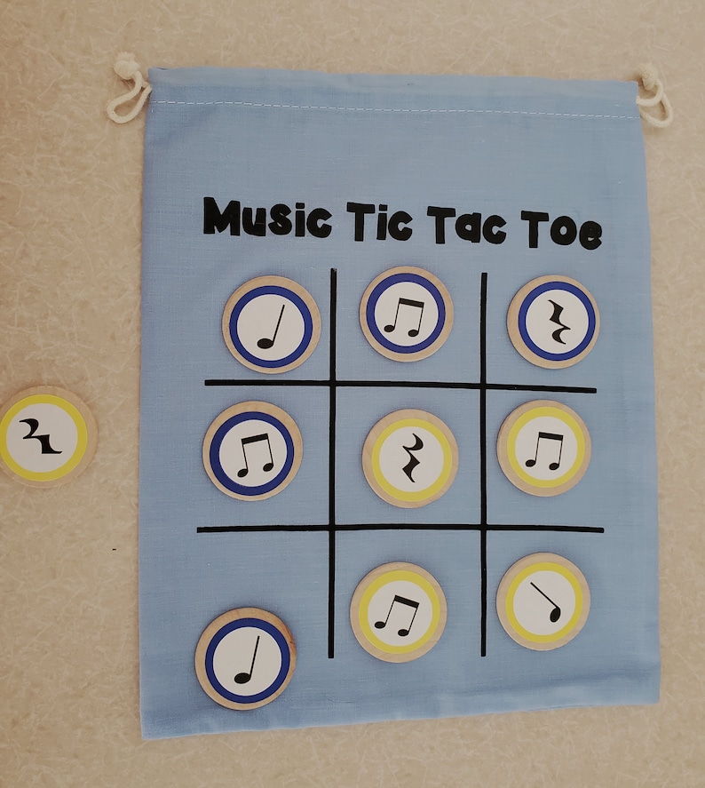 Puede incluir: Un tablero de juego de tela azul claro con el texto "Music Tic Tac Toe" y una cuadr&iacute;cula para jugar. Las fichas de madera presentan notas musicales. Una bolsa con cord&oacute;n est&aacute; adjunta en la parte superior.
