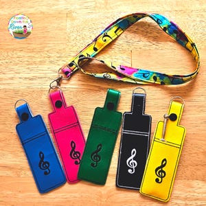 May include: Five colorful keychains with a treble clef design. Each keychain is made of a different color fabric: blue, pink, green, black, and yellow. The keychains are attached to a lanyard with a musical note pattern.