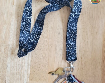 May include: A black lanyard with white musical notes printed on it. The lanyard has a silver key ring with keys and a small charm attached to it.
