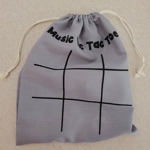 Puede incluir: Una bolsa gris claro con cord&oacute;n, con las palabras "Music Tic Tac Toe" impresas en negro. Una cuadr&iacute;cula de tres en raya negra est&aacute; impresa en la parte delantera. La bolsa tiene un cierre de cord&oacute;n blanco.