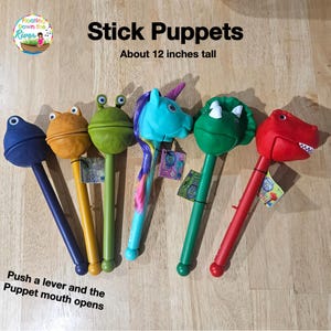 May include: A collection of six colorful stick puppets, each approximately 12 inches tall. The puppets include a blue blob, an orange frog, a green frog, a blue unicorn, a green dinosaur, and a red dinosaur. Text on the image reads "Stick Puppets" and "Push a lever and the Puppet mouth opens."