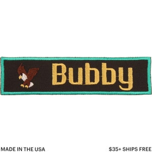 May include: Embroidered patch with the name "Bubby" in gold lettering on a black background. A brown and white eagle graphic is on the left. The patch has a teal border. Text at the bottom reads "MADE IN THE USA".