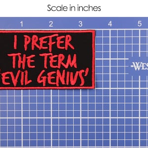 May include: Black rectangular patch with red embroidered text: "I PREFER THE TERM 'EVIL GENIUS'". The patch is approximately 4 inches wide and 2 inches tall. The patch is on a blue grid background with a ruler.