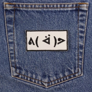 Pu&ograve; includere: Primo piano di una tasca di jeans in denim blu con una toppa rettangolare bianca. La toppa presenta simboli e caratteri neri. Il denim ha un aspetto classico e usurato con cuciture visibili.