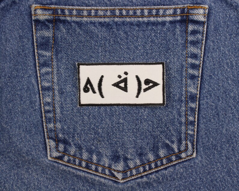 Pu&ograve; includere: Primo piano di una tasca di jeans in denim blu con una toppa rettangolare bianca. La toppa presenta simboli e caratteri neri. Il denim ha un aspetto classico e usurato con cuciture visibili.