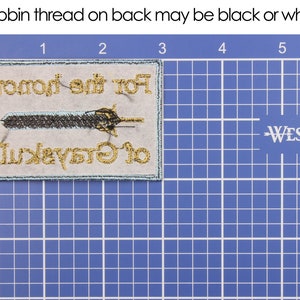 May include: Embroidered patch with the text "For the Honor of Greyskull" and a sword design. The patch has gold and black thread on a light gray background with a teal border. The bobbin thread on the back may be black or white.