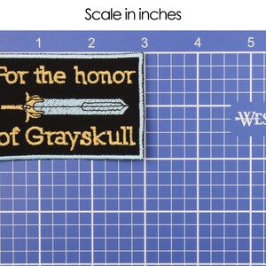 May include: Black rectangular patch with gold embroidered text reading "For the honor of Grayskull." A light blue sword design is centered on the patch. The patch is approximately 2 inches by 3 inches.