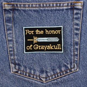 May include: A rectangular black patch with gold embroidered text that reads "For the honor of Grayskull" and a light blue sword design. The patch is sewn onto the back pocket of a blue denim jean.
