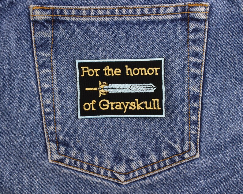 May include: A rectangular black patch with gold embroidered text that reads "For the honor of Grayskull" and a light blue sword design. The patch is sewn onto the back pocket of a blue denim jean.