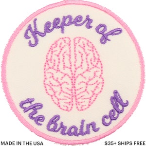 Può includere: Toppa rotonda ricamata con sfondo color crema, bordo rosa e disegno di cervello rosa. Il testo "Keeper of the brain cell" è ricamato con filo viola. La toppa ha un aspetto cucito ed è adatta per abbigliamento o borse.