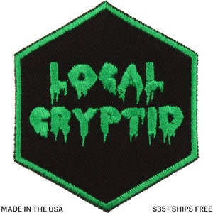 May include: A black, hexagonal patch with green embroidered text that reads "LOCAL CRYPTID." The text has a dripping effect. The patch has a green border and the words "MADE IN THE USA" are at the bottom.