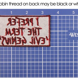 May include: A rectangular patch with the text "I Prefer The Devil I Know" in red, stitched onto a light gray fabric. The patch has a black border and is placed on a blue grid background.