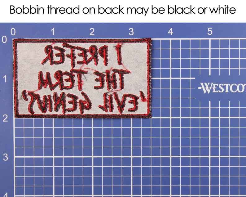 May include: A rectangular patch with the text "I Prefer The Devil I Know" in red, stitched onto a light gray fabric. The patch has a black border and is placed on a blue grid background.