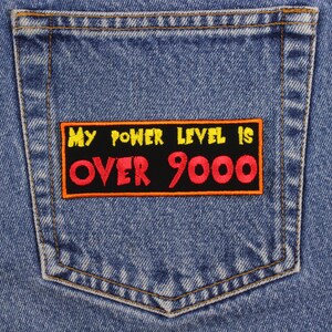 May include: Close-up of a light blue denim jeans pocket with a rectangular patch. The patch has the text "MY POWER LEVEL IS OVER 9000" in yellow and red lettering on a black background. The patch is outlined in orange.