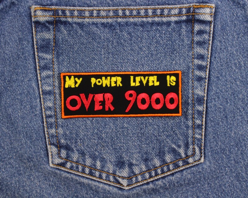 May include: Close-up of a light blue denim jeans pocket with a rectangular patch. The patch has the text "MY POWER LEVEL IS OVER 9000" in yellow and red lettering on a black background. The patch is outlined in orange.