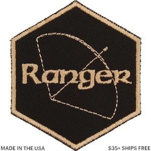 Puede incluir: Parche bordado negro y dorado con la palabra "Ranger" y un diseño de arco y flecha. El parche tiene forma hexagonal con un borde dorado. El texto "MADE IN THE USA" está en la parte inferior.