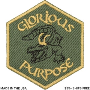 May include: A green, hexagonal patch with gold trim and the words "GLORIOUS PURPOSE" in gold lettering. A black and green alligator graphic is in the center. The patch is made in the USA.