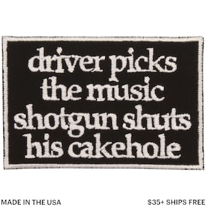 Op de afbeelding: Zwart rechthoekig embleem met witte geborduurde tekst: "driver picks the music shotgun shuts his cakehole". De patch is omrand met witte stiksels en bevat de tekst "MADE IN THE USA".