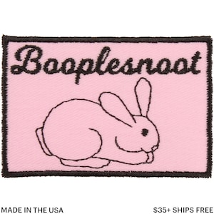 Puede incluir: Un parche rectangular rosa con la palabra "Booplesnoot" bordada en escritura negra sobre un dibujo lineal de un conejo. El conejo también está delineado en negro. El parche tiene un borde negro.