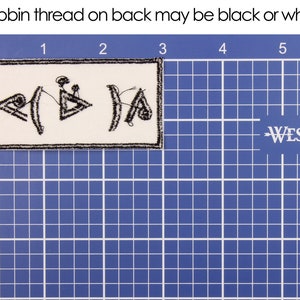 Pu&ograve; includere: Una toppa rettangolare con simboli ricamati neri su sfondo bianco. La toppa &egrave; bordata con cuciture nere. Il testo "Bobbin thread on back may be black or white" &egrave; sopra la toppa. La toppa &egrave; su una griglia blu.