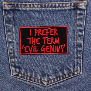 May include: Close-up of a blue denim jeans pocket with a black rectangular patch. The patch has red text that reads "I PREFER THE TERM 'EVIL GENIUS'". The patch is sewn onto the pocket.