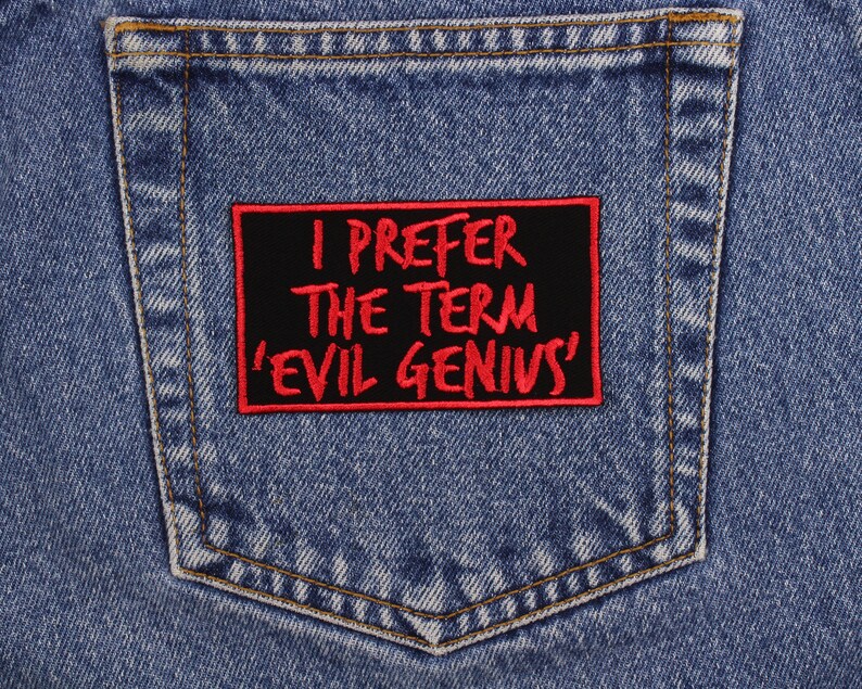 May include: Close-up of a blue denim jeans pocket with a black rectangular patch. The patch has red text that reads "I PREFER THE TERM 'EVIL GENIUS'". The patch is sewn onto the pocket.