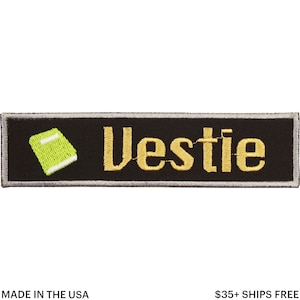 May include: Black rectangular patch with the word "Vestie" embroidered in gold, accompanied by a small green book icon. The patch has a silver border and is labeled "Made in the USA".