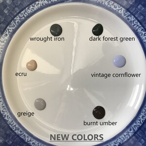 May include: A white plate with six paint samples in different colors. The colors are: wrought iron, dark forest green, vintage cornflower, ecru, greige, and burnt umber. The plate has a blue and white patterned border. The text "NEW COLORS" is printed below the plate.
