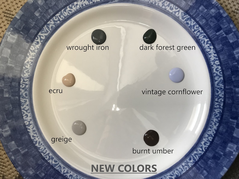 May include: A white plate with six paint samples in different colors. The colors are: wrought iron, dark forest green, vintage cornflower, ecru, greige, and burnt umber. The plate has a blue and white patterned border. The text "NEW COLORS" is printed below the plate.