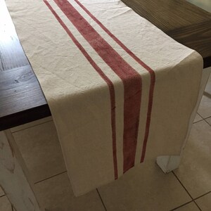 May include: A white linen table runner with three wide red stripes. The runner is laying on a wooden table.