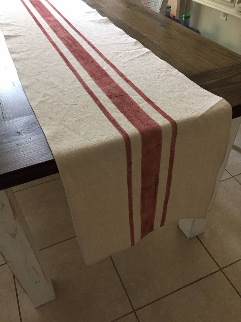 May include: A white linen table runner with three wide red stripes. The runner is laying on a wooden table.
