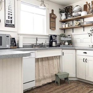 May include: A white kitchen with white cabinets, a stainless steel sink, and a white dishwasher. There is a beige and brown striped curtain hanging under the sink. A small green stool sits in front of the curtain. There is a brown rug on the floor. The kitchen has a window with a white frame and a white countertop. There are shelves on the wall with various items on them, including a coffee maker, a wooden cutting board, and a white pitcher. The shelves are made of wood and have black metal brackets. There is a sign on the wall that says "Kitchen Conversions".
