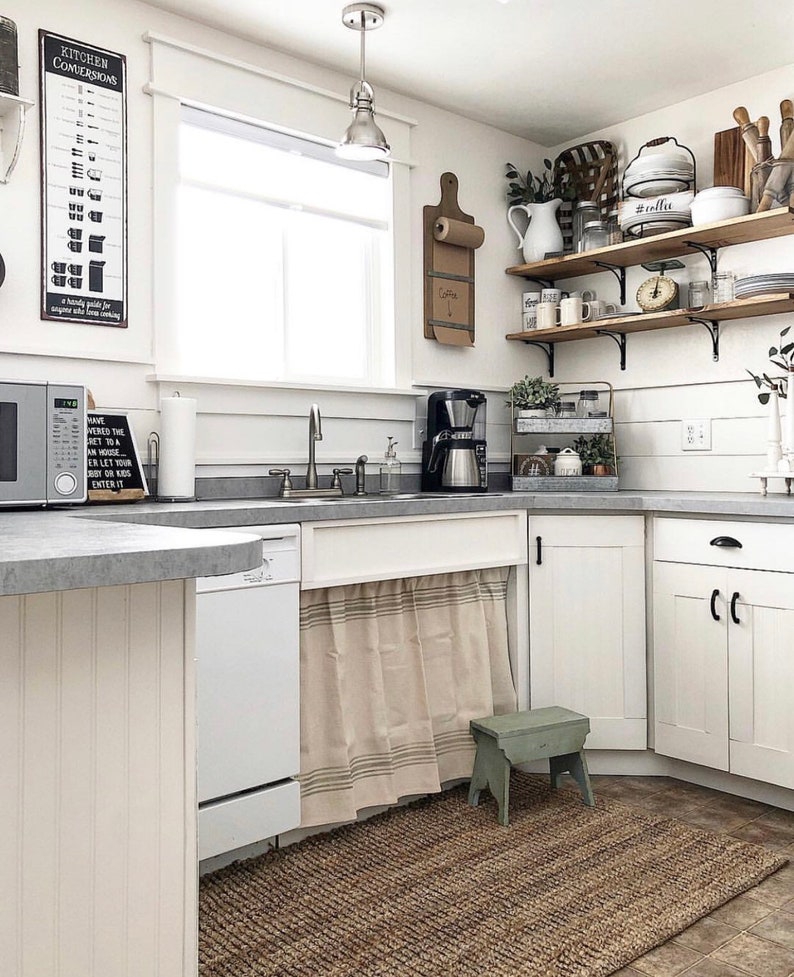 May include: A white kitchen with white cabinets, a stainless steel sink, and a white dishwasher. There is a beige and brown striped curtain hanging under the sink. A small green stool sits in front of the curtain. There is a brown rug on the floor. The kitchen has a window with a white frame and a white countertop. There are shelves on the wall with various items on them, including a coffee maker, a wooden cutting board, and a white pitcher. The shelves are made of wood and have black metal brackets. There is a sign on the wall that says "Kitchen Conversions".