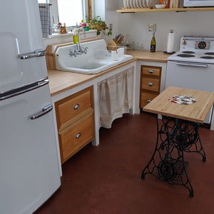 May include: A white vintage refrigerator with a double door, a white farmhouse sink with a chrome faucet, and a wooden countertop with drawers. The countertop is also used as a table with a black metal base.