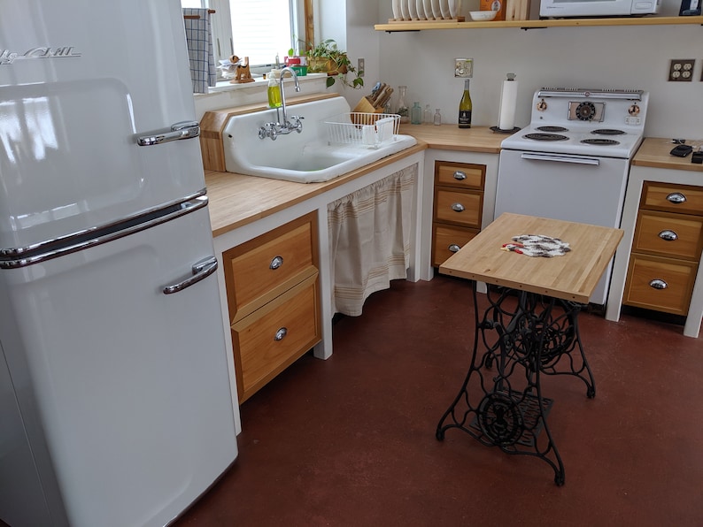 May include: A white vintage refrigerator with a double door, a white farmhouse sink with a chrome faucet, and a wooden countertop with drawers. The countertop is also used as a table with a black metal base.