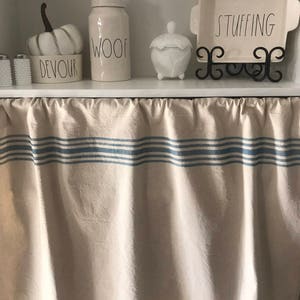 May include: A white kitchen shelf with a white pumpkin, a white jar labeled "WOOF", a white ceramic jar with a lid, and a white baking dish on a black stand labeled "STUFFING". The shelf has a white valance with blue stripes.