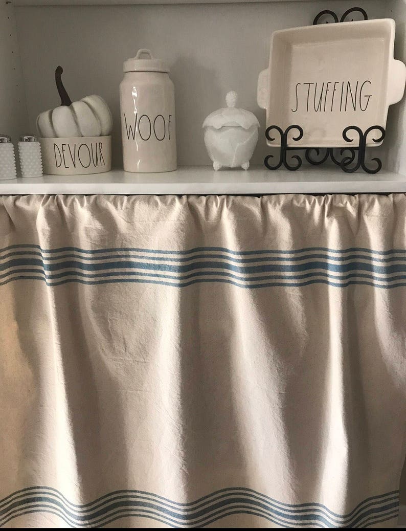 May include: A white kitchen shelf with a white pumpkin, a white jar labeled "WOOF", a white ceramic jar with a lid, and a white baking dish on a black stand labeled "STUFFING". The shelf has a white valance with blue stripes.