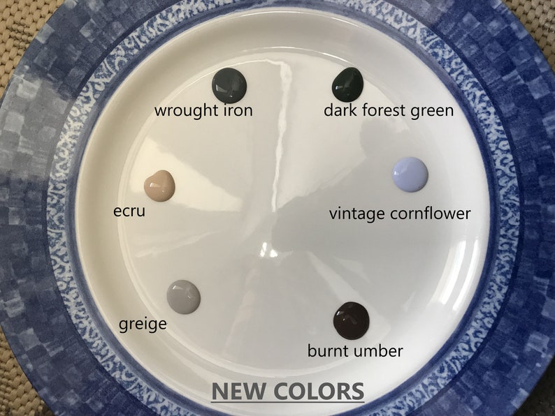 May include: A white plate with six paint samples in different colors. The colors are: wrought iron, dark forest green, vintage cornflower, ecru, greige, and burnt umber. The plate has a blue and white patterned border. The text "NEW COLORS" is printed below the plate.