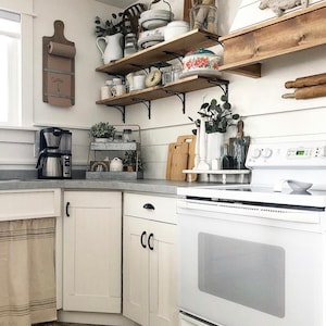 May include: White kitchen with white cabinets, a white stove, and wooden shelves with various kitchen items. The shelves are mounted on the wall and are filled with dishes, jars, and other kitchen supplies. There is a wooden cutting board on the counter and a coffee maker on the counter. The text "Coffee" is written on a piece of paper on the wall.