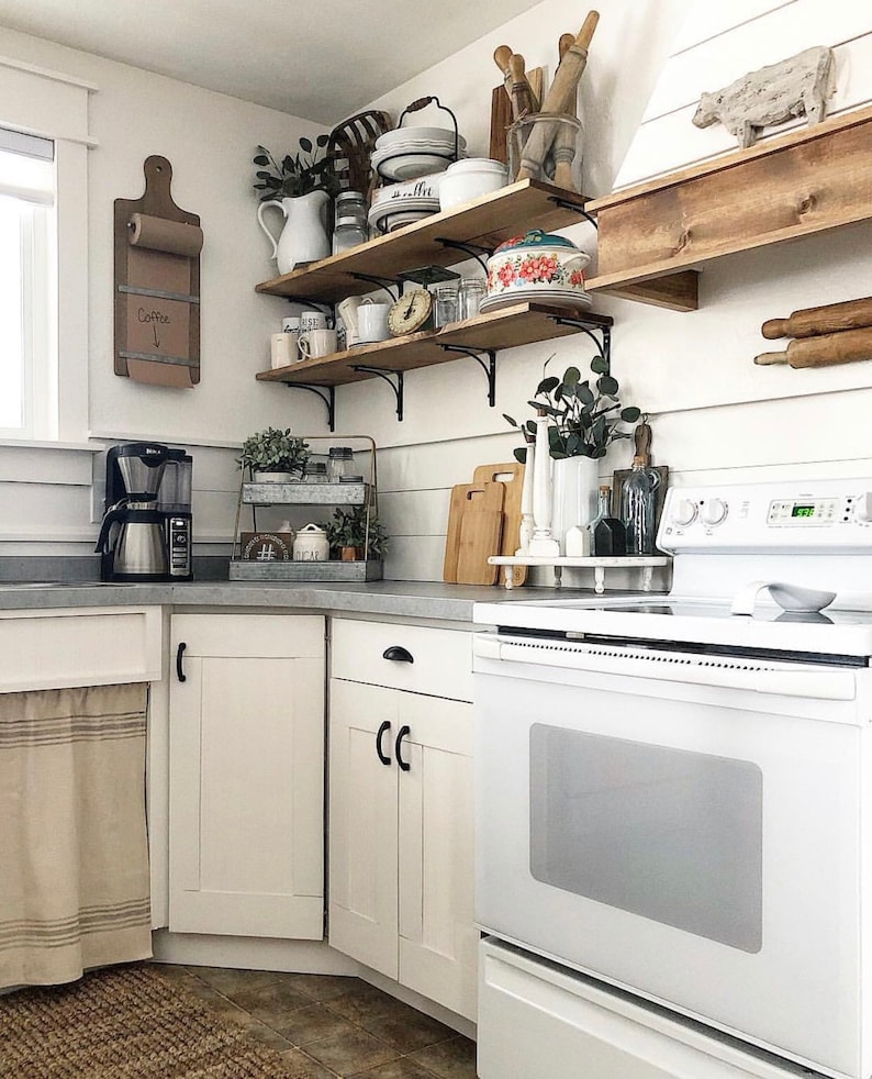 May include: White kitchen with white cabinets, a white stove, and wooden shelves with various kitchen items. The shelves are mounted on the wall and are filled with dishes, jars, and other kitchen supplies. There is a wooden cutting board on the counter and a coffee maker on the counter. The text "Coffee" is written on a piece of paper on the wall.