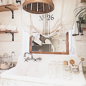 May include: A white farmhouse style kitchen sink with a chrome faucet and a wooden basket hanging above. The sink has a white countertop and a white tiled backsplash. There are several glass jars and a glass pitcher on the counter. The basket has the number "26" printed on it.
